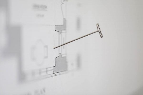 Close-up of a T-shaped pin inserted into a grayscale architectural floor plan, displayed vertically on a wall. The plan includes various lines and shapes suggestive of rooms and structural elements, with blurred text and details in the background.