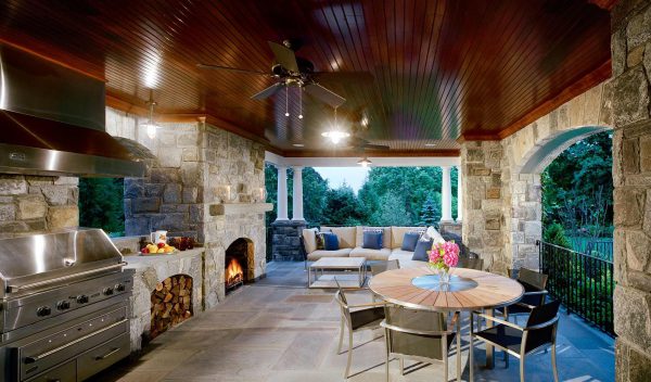 Outdoor patio with stone walls and arched openings. Features a stainless steel grill, wood-burning fireplace, and a ceiling fan on a wooden ceiling. Seating area includes a cozy sectional sofa and a round dining table with four chairs. A view of lush greenery is visible outside.