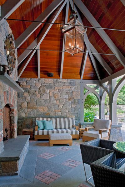 A cozy outdoor patio features a pitched wooden ceiling and stone walls. A striped sofa with cushions, a wicker chair, and a table with wicker chairs provide seating. A large lantern-style light hangs from above, and a brick fireplace adds warmth. Greenery is visible outside.