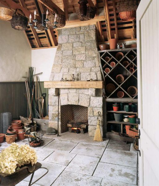 A rustic room with a large stone fireplace. Firewood is burning inside the hearth. Surrounding the fireplace are shelves with potted plants and garden tools. The ceiling has hanging baskets and a chandelier. A broom leans against the fireplace. Cream-colored door on the right.