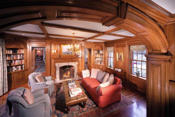 Luxurious wood-paneled living room with intricate ceiling beams. Features a red sofa, two blue armchairs, and a wooden coffee table on a patterned rug. A lit fireplace is flanked by built-in bookshelves. An archway leads to a dining room, and windows have floral valances. A chandelier hangs above.