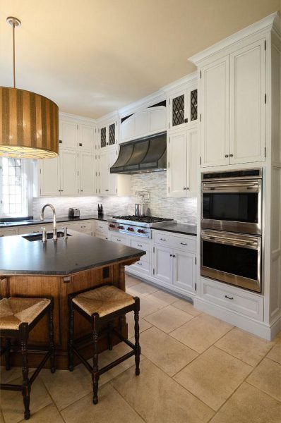 A spacious kitchen with white cabinetry and a large island. The island has a dark countertop and three wooden stools. Stainless steel appliances, including a double oven and range, are built into the cabinets. The backsplash features gray tiles, and a large brown pendant light hangs above.