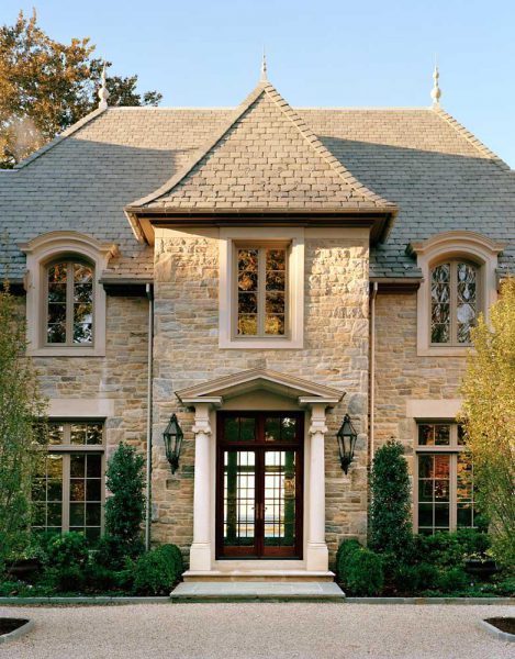 A large stone house with a steep gray slate roof and pointed tower-like structures. The front features three tall windows and a dark wooden door with glass panels, framed by white columns. Two symmetrical, lantern-style lights are mounted beside the entrance. Neat shrubbery lines the walkway.