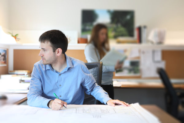 A man in a blue shirt sits at a desk, reviewing architectural plans with a pencil. Behind him, a woman stands reading a book. The office is bright, with a corkboard and various documents in the background. The scene suggests a collaborative office environment.