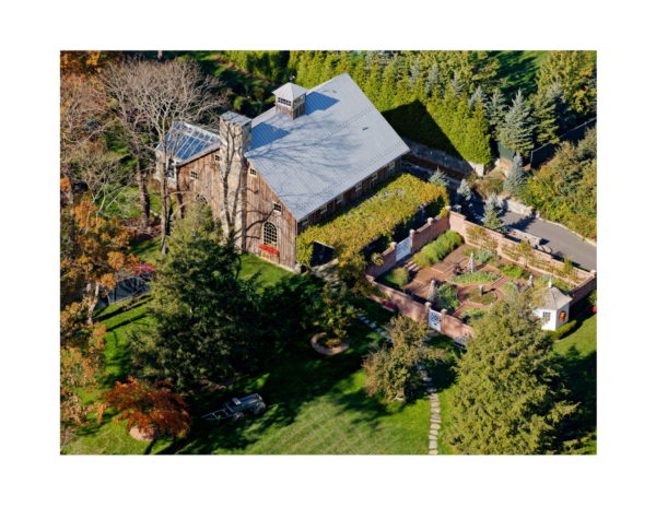 Aerial view of a large rustic barn with a metal roof surrounded by dense trees. This charming outdoor space includes a formal garden featuring a circular pathway and brick walls. The landscape is lush with green grass and diverse trees, merging evergreens with deciduous highlights showcasing springs vibrance.