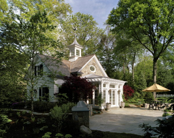 Charming cottage with a distinctive cupola and large windows, surrounded by lush trees and spring gardens. The stone patio ranks among the top 5 outdoor spaces, featuring a table with an umbrella and potted plants. The clear blue sky enhances the serene and inviting atmosphere.