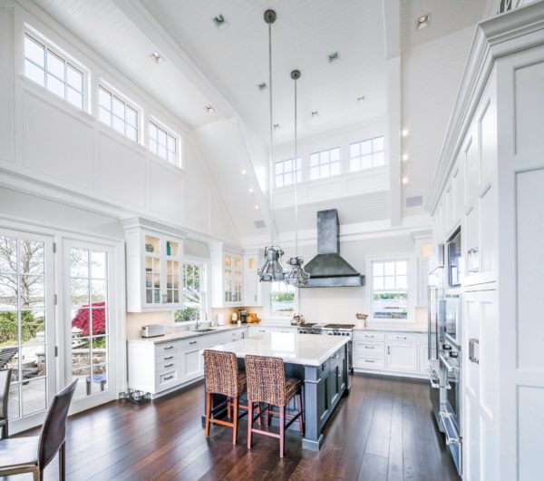 Spacious kitchen with high vaulted ceiling and large windows. Central island with white countertop and two wicker chairs showcase requested features in modern kitchen design. Black range hood above a stove, surrounded by white cabinetry. Dark hardwood floors contrast with white walls. Patio doors on the left.