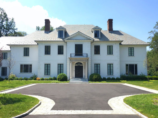 A large, two-story white mansion with a gray slate roof and two prominent chimneys showcases an impressive architectural design. It features a central columned entrance adorned with a small balcony above. Surrounded by green lawns and small trees, the property includes a circular driveway in the foreground.