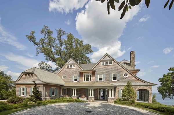 A large, elegant seaside house with a shingle exterior is nestled among mature trees. The home features multiple peaked roofs and a prominent stone chimney. In front is a circular driveway paved with cobblestones. The sky is partly cloudy, and the property overlooks a body of water.