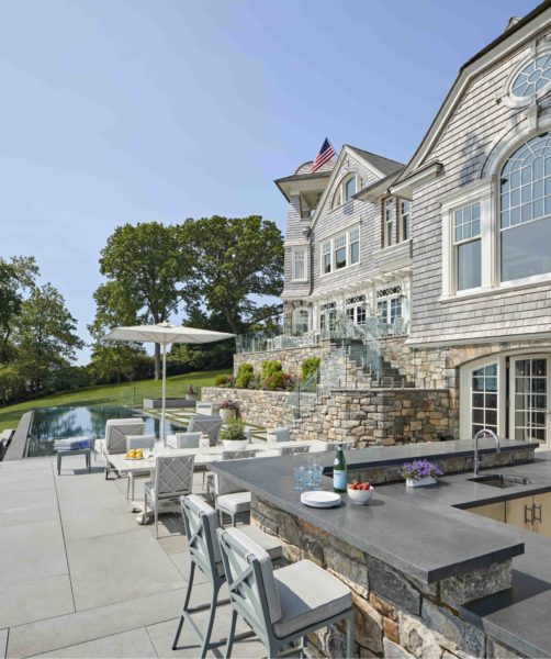 Luxurious outdoor space with a stone patio, bar, and stools overlooking an infinity pool. The backdrop features a large, traditional-style mansion with gray shingle siding and a turret. The scene includes a bottle of wine, glasses on the bar, and scenic views of lush greenery and clear sky.