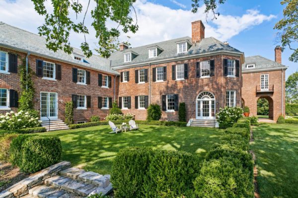 A large, elegant red-brick mansion with white-trimmed windows, black shutters, and chimneys. The house features a central, arched doorway and multiple dormer windows. The manicured lawn has hedges, a stone pathway, and two white Adirondack chairs. The sky is partly cloudy.