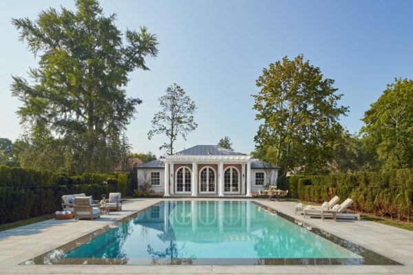 A serene outdoor scene features a rectangular swimming pool reflecting the clear blue sky, surrounded by patio tables and cushioned chairs. In the background is a classic white pool house with arched doors and windows, flanked by tall trees and lush greenery.