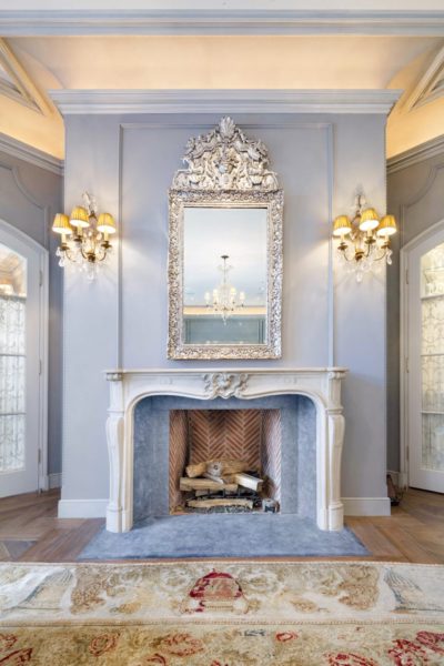 Elegant living room featuring sophisticated fireplace designs, with a marble fireplace topped by an ornate silver mirror. Vintage wall sconces with lampshades flank the mirror on light blue walls. A chandelier gleams in the reflection, while a patterned rug adorns the wooden floor.