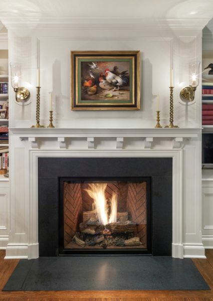 A fireplace with a black mantel and brick interior sets the tone for sophisticated home decor. Above it, a painting of chickens and fruit in a gold frame adds charm, flanked by brass wall sconces. Built-in shelves showcase books and decor items, enhancing the elegant design.