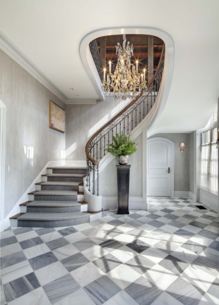 The grand foyer design showcases a sweeping staircase with a wooden handrail and ornate iron balustrade. A crystal chandelier casts light on the checkered marble floor, where a tall potted plant stands on a black pedestal. A large window and decorative painting complete this welcoming entry.