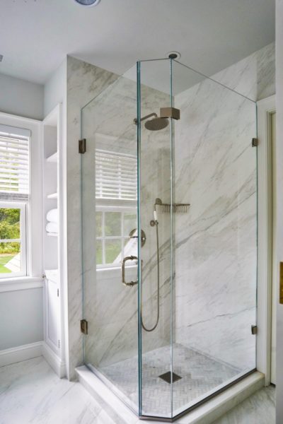 Modern bathroom with a glass-enclosed shower, featuring a rainfall showerhead. Renovation includes walls and floor lined with white marble tiles. Two windows with white blinds provide natural light. White towels are neatly stacked on shelves in this charming Colonial Pool Cottage setting.