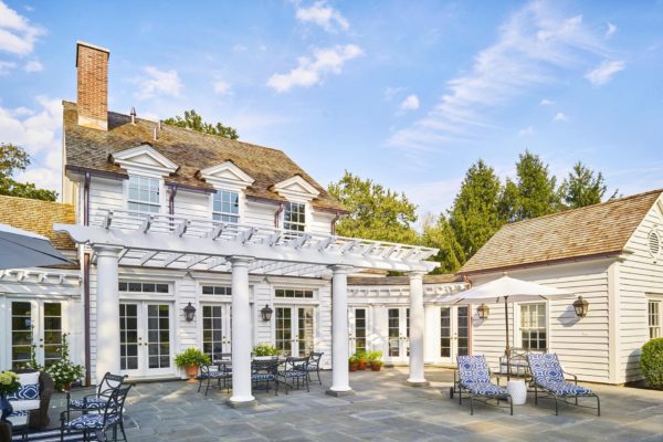 A charming Colonial two-story white house features a patio with a pergola, wicker chairs, and cushioned loungers. Black furniture and potted plants add elegance. The sky is clear with few clouds, while tall trees surround the property, enhancing its serene ambiance—a perfect home tour destination.
