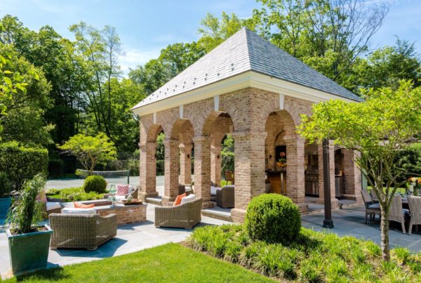 The outdoor seating area boasts a brick pavilion with an arched design reminiscent of Alice Washburn Awards architecture. Comfortable wicker chairs with cushions are arranged around a small table, surrounded by lush greenery and manicured shrubs, with a tennis court visible against the clear blue sky.