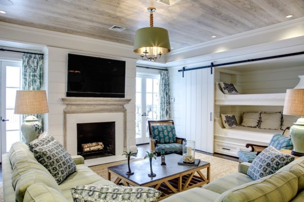 A stylish living room with a green and beige color scheme features a fireplace topped by a flat-screen TV, surrounded by sleek green sofas and armchairs. A rustic wooden coffee table sits at the center, while built-in bunk beds with curtains add charm on the right. A ceiling lamp casts a warm glow from above.