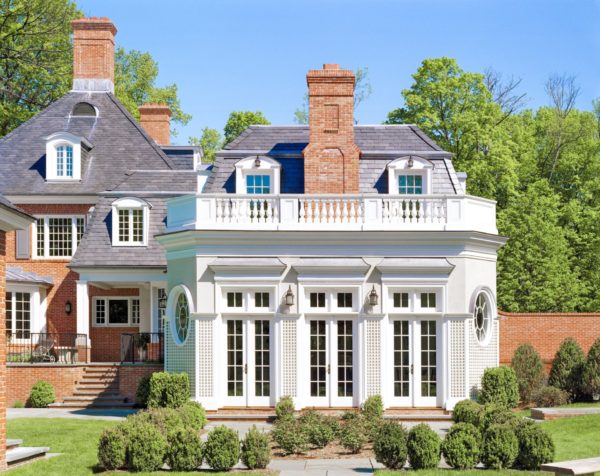 A large, elegant brick home designed by a renowned architect with a slate roof and multiple chimneys is shown. Nestled in the suburbs, it features tall, arched windows and a decorative balustrade. Surrounded by lush greenery under a clear blue sky, it conveys luxury and tranquility.