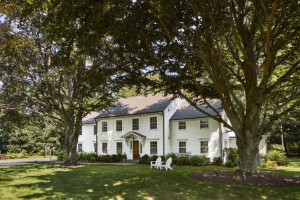 Relocating to the suburbs unveils gems like this large white colonial-style house with multiple windows and a shingled roof, nestled in lush greenery. Mature trees frame the scene while white Adirondack chairs adorn the sunlit lawn, inviting you to find tranquility amid natures embrace.
