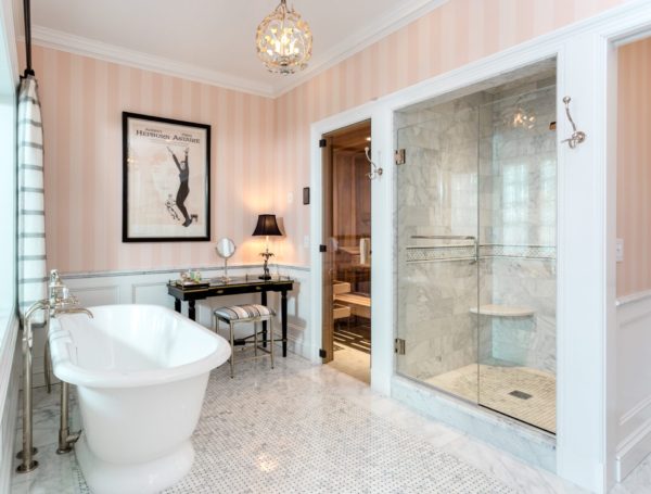 Elegant bathroom with a freestanding bathtub on a mosaic tile floor. Marble shower with glass door to the right. Black vanity table with a chair and lamp. A framed vintage poster graces the peach striped wall. Two small chandeliers hang from the ceiling, enhancing this luxury bath design as light streams in through a window.