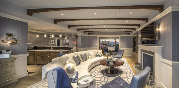 Elegant living room with a curved white sofa facing a fireplace, flanked by armchairs on a patterned rug. The ceiling has wooden beams and recessed lighting. A bar area with pendant lights is visible, and a large window offers a glimpse of an outdoor scene.