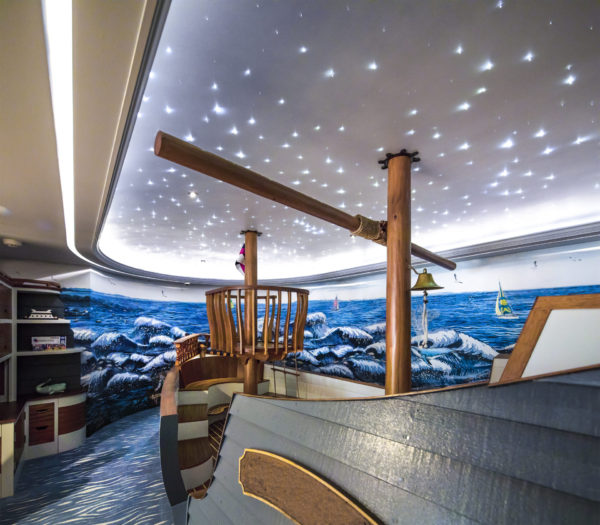 Room with a ship-themed design. A wooden ship structure with a mast and crows nest occupies the space. The walls feature a mural of ocean waves with small boats, and the ceiling resembles a starry night sky, with small lights resembling stars. A bell hangs nearby.