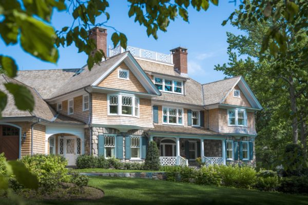 A large, elegant house with blue shutters and a mix of stone and wood exterior exudes classic Shingle Style architecture amidst lush green trees. It features multiple chimneys, gabled roofs, and white railings. The well-maintained lawn enhances its charm under a bright blue sky.