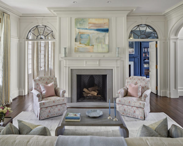 Elegant living room with a central fireplace flanked by two arched windows. Above the fireplace hangs an abstract painting. Two patterned armchairs with pink cushions face a dark coffee table. Soft neutral tones dominate, with curtains framing the windows and a glimpse of a blue room beyond.