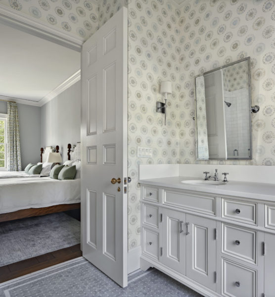 Elegant bathroom with a white vanity, sink, and wall-mounted mirror. Soft-patterned wallpaper featuring circular designs. Doorway leads to a bedroom with a bed, green pillows, and patterned curtains. Natural light from a window illuminates both spaces.