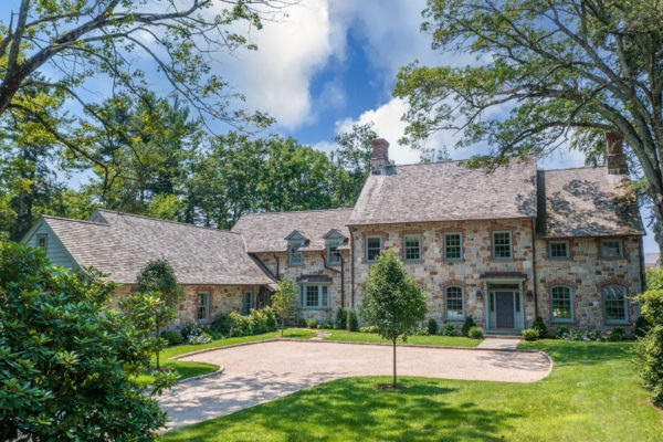 A large stone house with a gabled roof, surrounded by lush greenery and tall trees. The facade features multiple windows with shutters and a central doorway. A circular gravel driveway is in front, edged by manicured grass. The sky is partly cloudy with ample sunlight.