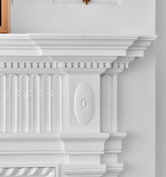Close-up of a white, intricately designed fireplace. Features embossed vertical ovals, each with a small central dot. A central medallion with radiating lines adds texture. The mantelpiece has layered moldings, creating a classic and ornate architectural look.