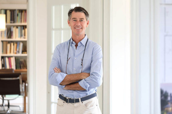 A middle-aged man with gray hair stands confidently with arms crossed. He wears a light blue shirt and beige pants with a woven belt. Glasses hang from his neckline. The background features bookshelves and modern office furniture with soft lighting.