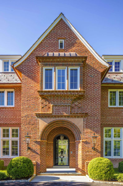 A brick house featuring a gabled roof and an arched doorway. The facade has symmetrical windows with white trim and decorative brickwork. Two lantern-style lights flank the entrance. Well-manicured shrubs are positioned on either side of the path leading to the door.