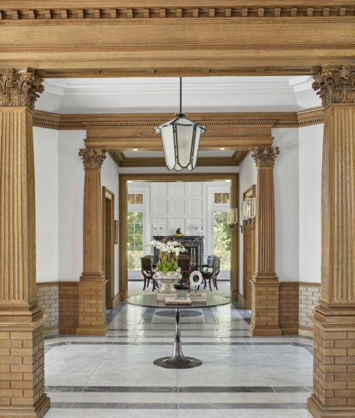 Ornate hallway with fluted wooden columns and brick bases. A round table with books and flowers is centered on a tile floor. Beyond the table, a dining area with dark wood furniture is visible. A geometric pendant light hangs from a coffered wooden ceiling. Large windows let in natural light.