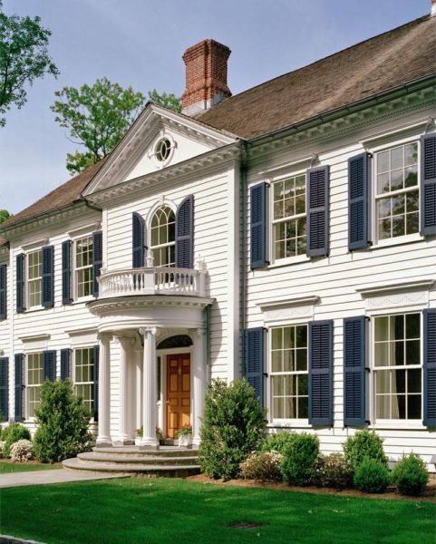 A large, two-story white colonial house with dark blue shutters and a red brick chimney. It features a prominent arched window above a rounded balcony supported by columns over the wooden front door. Lush green lawn and shrubs surround the house under a clear blue sky.