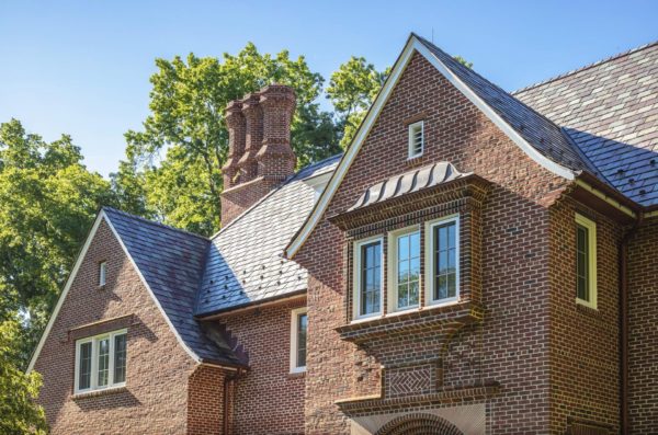 A brick house with a steep, slate roof features a prominent brick chimney with decorative elements. The house has multiple windows with white frames and panes. Lush green trees are visible in the background against a clear blue sky, indicating a sunny day.