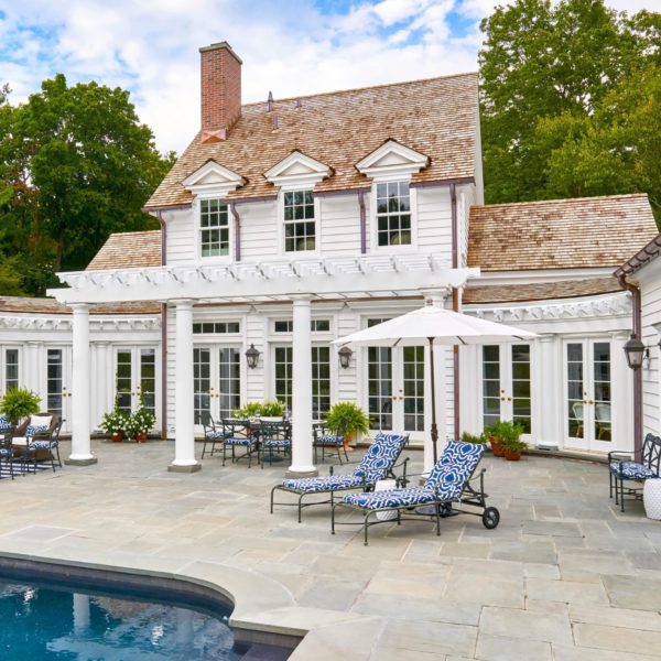 White colonial-style house with a shingle roof, featuring tall windows and a central chimney. A stone patio surrounds a pool, with blue patterned loungers under an umbrella and dining table nearby. Greenery decorates the space. Trees are visible in the background, under a partly cloudy sky.