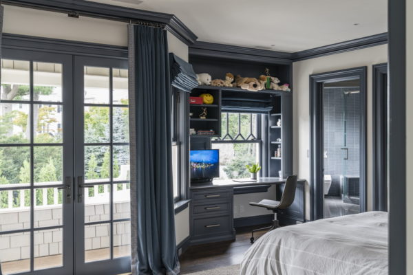 Bedroom with large glass doors leading to a balcony, blue-gray walls and furniture. A desk with a computer and chair is set against a window. Shelves above hold stuffed animals. The bed has white bedding. A vase with flowers is on the desk. A doorway leads to another room.