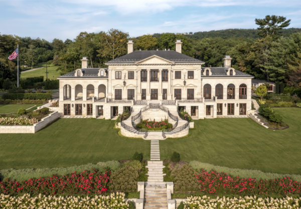 A large, elegant mansion with neoclassical architecture sits on a lush lawn. The symmetrical building features multiple columns, a dark roof, and numerous windows. A grand staircase leads to the entrance, surrounded by vibrant flowerbeds and manicured gardens, with a U.S. flag flying nearby.