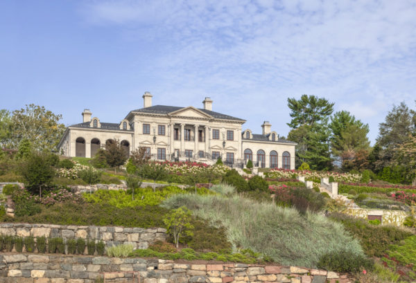 A large, elegant mansion sits atop a landscaped hill. The building features a neoclassical design with symmetrical columns and a grand facade. The lush gardens in the foreground include a variety of shrubs, flowers, and stone terraces. The sky is partly cloudy with blue hues.