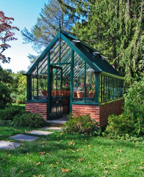 A small greenhouse made of glass with a red brick base stands in a lush garden. It features potted plants inside, visible through the clear panels. The surrounding area is green with trees and bushes, and sunlight casts gentle shadows on the scene, highlighting the vibrant foliage.