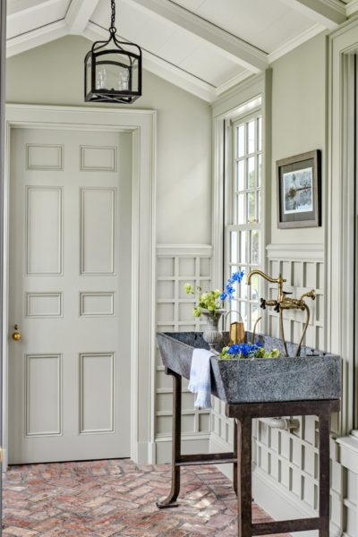 A rustic hallway with a brick floor features a large stone sink on a metal stand. Fresh wildflowers in the sink add color. Light gray paneled walls and ceiling are complemented by a window with grid detailing. A black pendant light and framed artwork decorate the space.