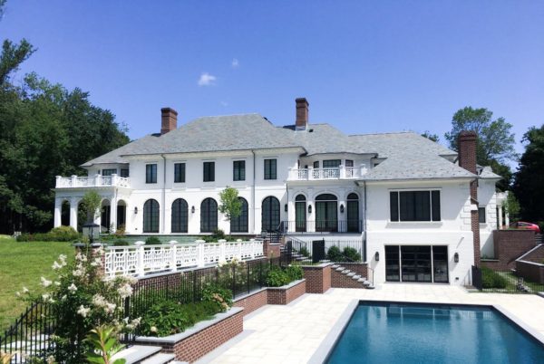 Luxurious two-story mansion showcasing Georgian architecture, with a gray slate roof, white exterior, and arched windows. The foreground features a large rectangular pool, landscaped garden beds, and brick accents. A spacious patio area with white railings overlooks the pool. Trees border the property.