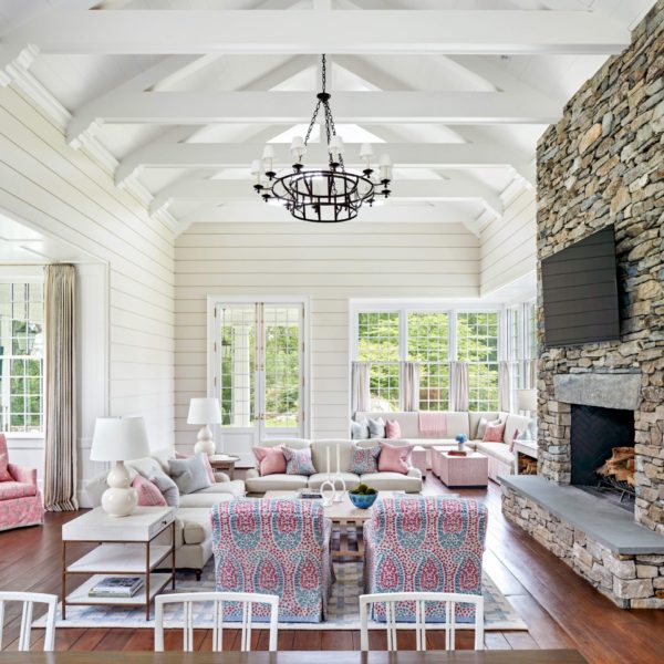 Spacious living room with white paneled walls, exposed beams, and a dark metal chandelier. This colonial home features a large stone fireplace, pink and white sofas, patterned armchairs, and a wooden floor. Large windows offer a view of greenery outside, adding natural light to the elegant decor.