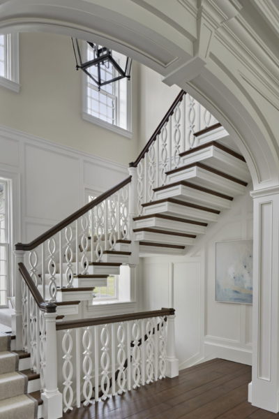 A white staircase with dark wood railing, featuring intricate cut-out balusters, curves upward through an arched entryway in this colonial home. The space is illuminated by a large, black lantern-style light fixture. A soft, abstract painting adorns the paneled wall as natural light streams through tall windows.