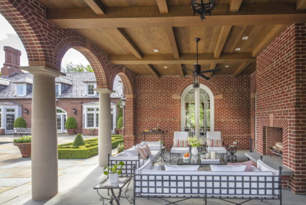 A spacious outdoor patio, perfect for enjoying serene outdoor spaces, features brick walls and a wooden ceiling with a white grid-patterned metal seating area. Soft-colored cushions adorn the seats. The patios arched openings and large glass door offer views of lush greenery in the background.