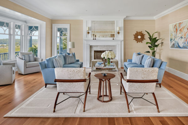 A spacious, bright living room with light wooden floors and beige walls. Blue sofas and patterned white chairs surround a wooden coffee table with a flower vase. Theres a large window with an outdoor view, a white fireplace, and a decorative mirror and plant.