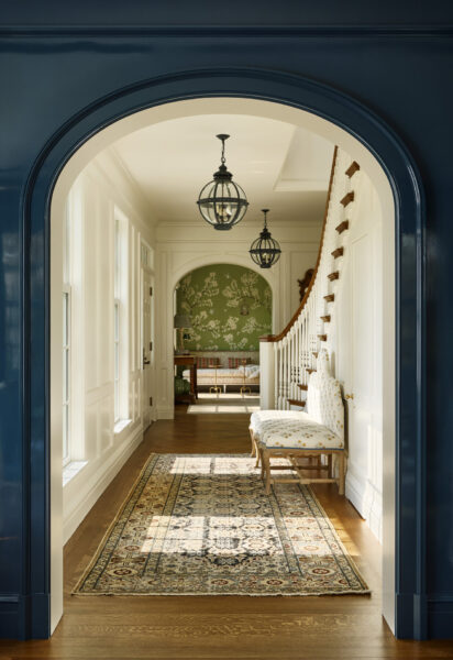 A hallway with a dark blue arched entrance, often highlighted in architecture on Instagram, features a staircase with a wooden handrail. A patterned runner rug leads to a room with floral wallpaper. Two lantern-style light fixtures hang from the ceiling, and a cushioned bench with patterned upholstery sits on the right.